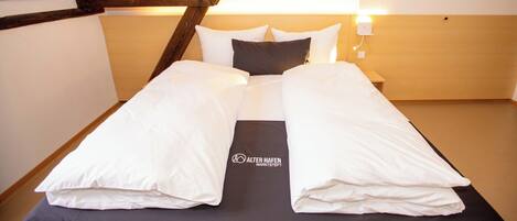Habitación de hotel con techo alto, vigas de madera, cama con ropa blanca y dos cuadros en la pared.