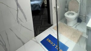 Une chambre d’hôtel avec un mur en marbre, un lit recouvert d’une courtepointe à rayures bleues et blanches, des toilettes blanches et un paillasson bleu.