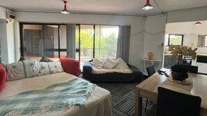 Une chambre d’hôtel comprenant un lit, un bureau, une chaise, une table à manger et un coin cuisine. Une grande fenêtre laisse entrer la lumière naturelle.
