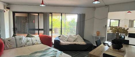 A hotel room with a bed, a desk, a chair, a dining table, and a kitchenette. There is a large window providing natural light.
