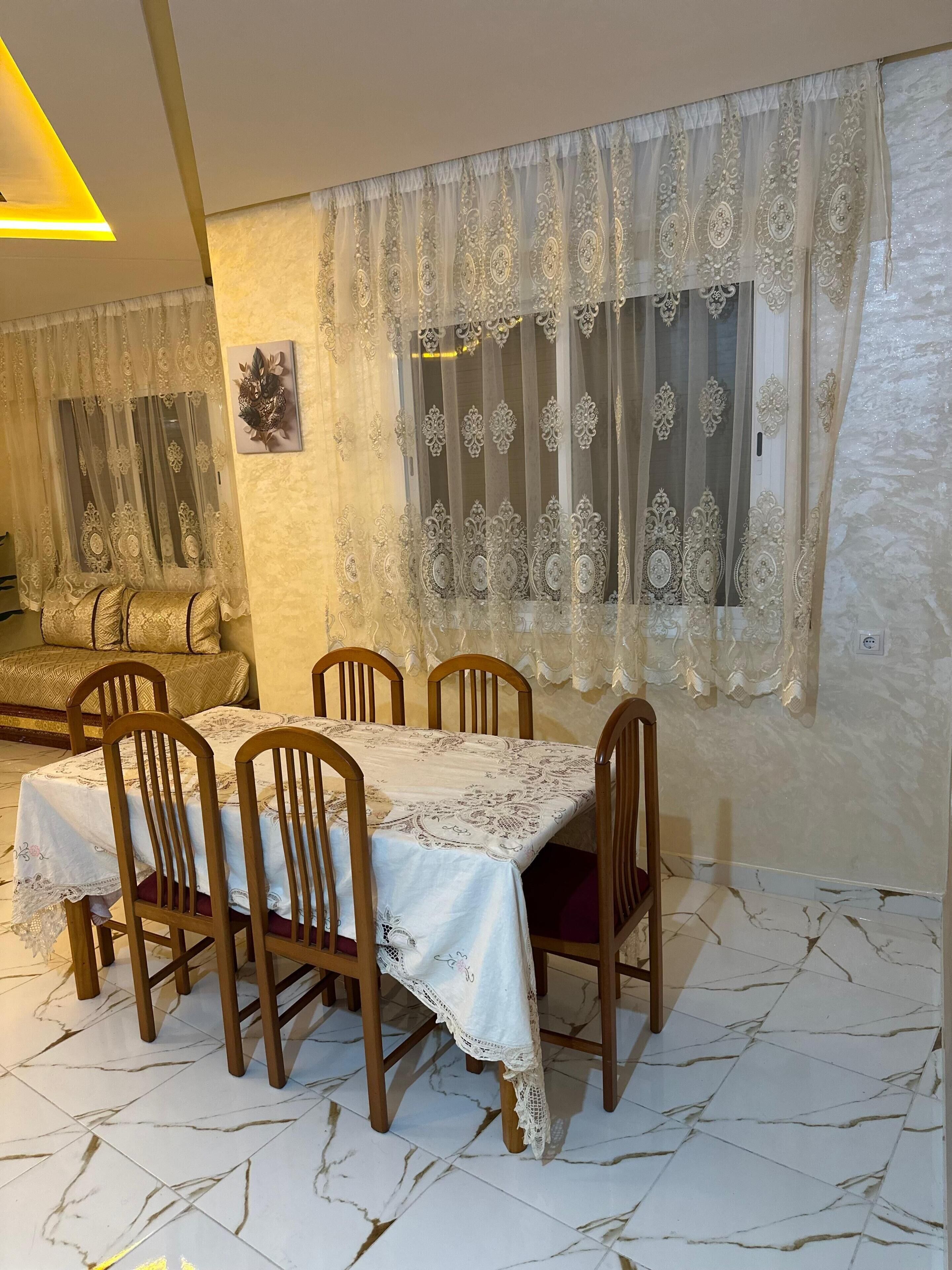 Un coin repas avec une table dressée pour quatre personnes, des chaises en bois, une nappe à motifs et une fenêtre agrémentée de rideaux décoratifs.