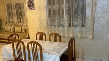 Un coin repas avec une table dressée pour quatre personnes, des chaises en bois, une nappe à motifs et une fenêtre agrémentée de rideaux décoratifs.