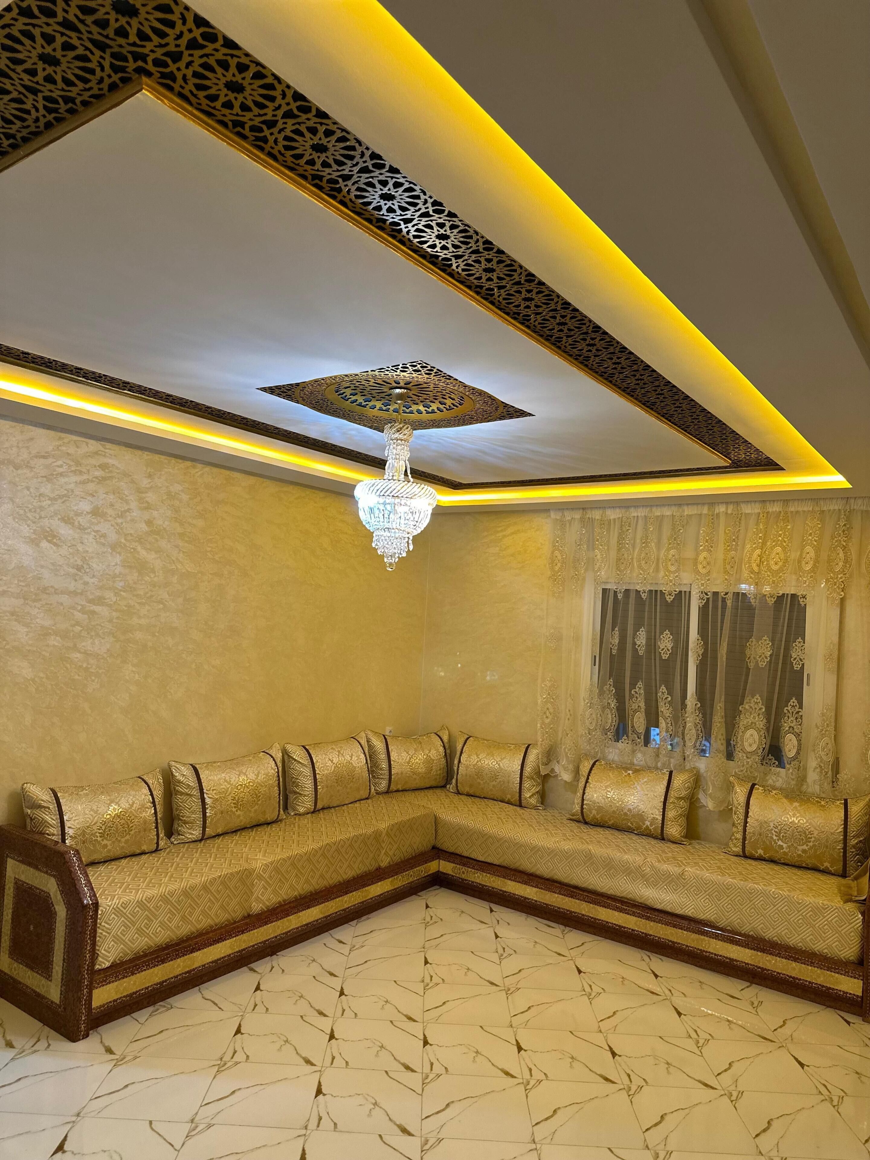 Une pièce avec un canapé d’angle, un lustre, un plafond décoré et une fenêtre avec des rideaux.