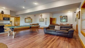 Ampla sala de estar com mesa de bilhar, sofá de couro, piso de madeira e cozinha ao fundo.