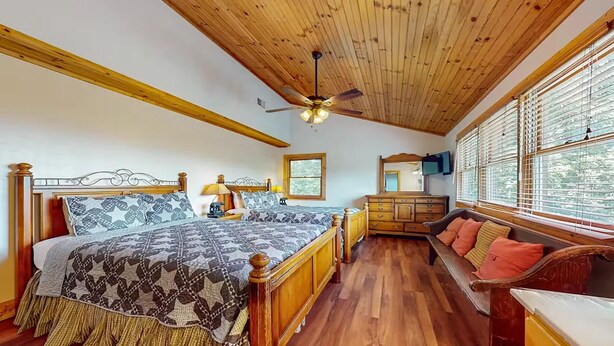 Quarto com teto de madeira, cama com roupa de cama estampada, cômoda, um banco com almofadas e uma televisão na parede.