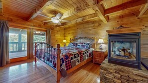 A wooden cabin bedroom with a bed, a stone fireplace, and a balcony. - Moonlit Mountain Retreat | Mountain Views, Hot Tub & Fantastic Game Room (Sevierville)