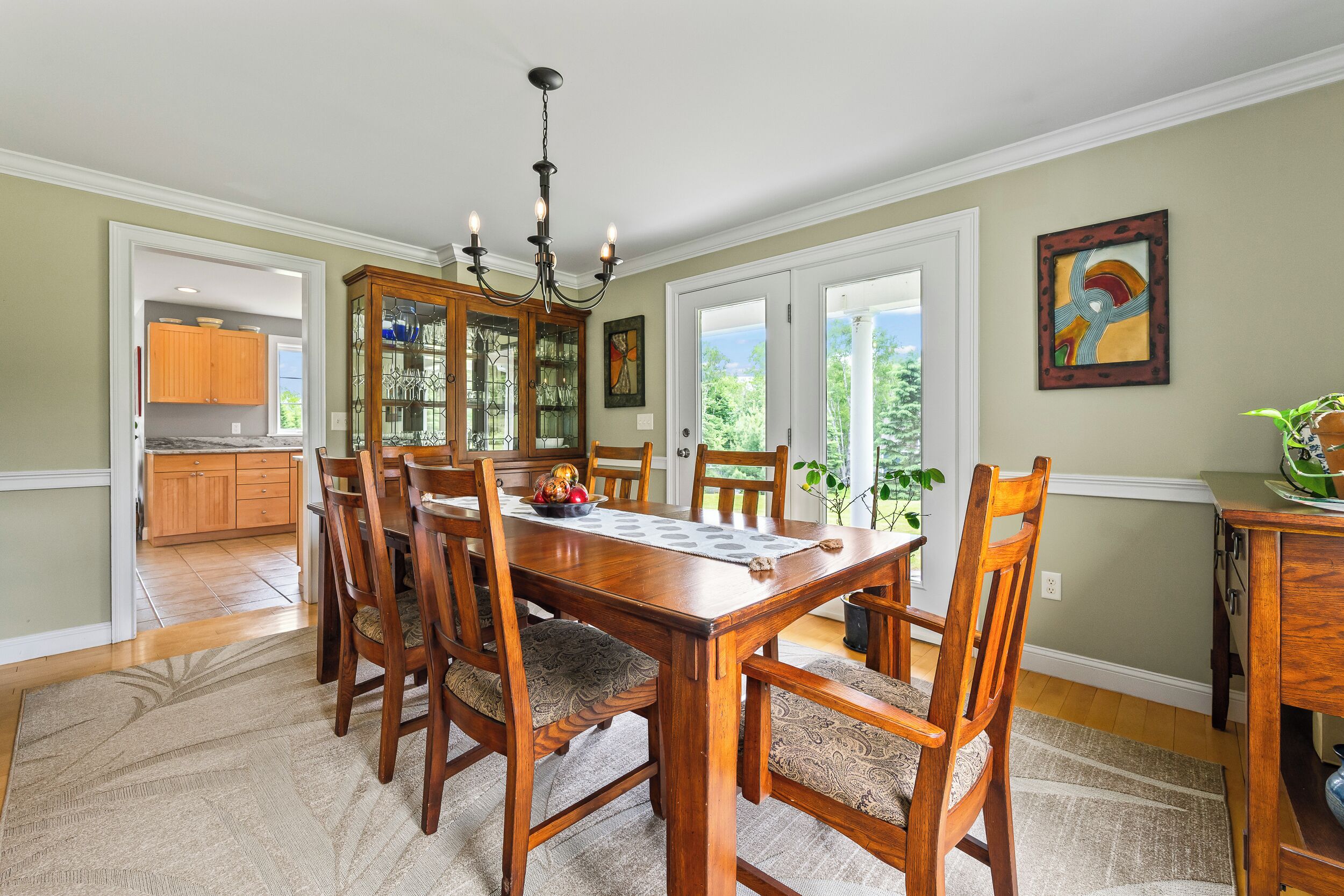 A dining room with a wooden table and chairs, a chandelier, and a view of the outdoors through large windows.