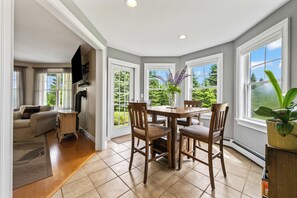 A spacious dining area with a wooden table and chairs, large windows offering a view of greenery, and a modern kitchen with a built-in oven. - Mingo Manor | Across from Mingo Springs Golf Course!! (Rangeley)