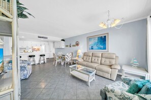 Ein modernes Wohnzimmer mit einer Couch, einem Couchtisch und Sesseln. Es gibt eine Küchenzeile mit weißen Schränken und einem Esstisch. Ein Gemälde hängt an der Wand, und auf einem Beistelltisch steht ein kleines Modell eines Segelboots.