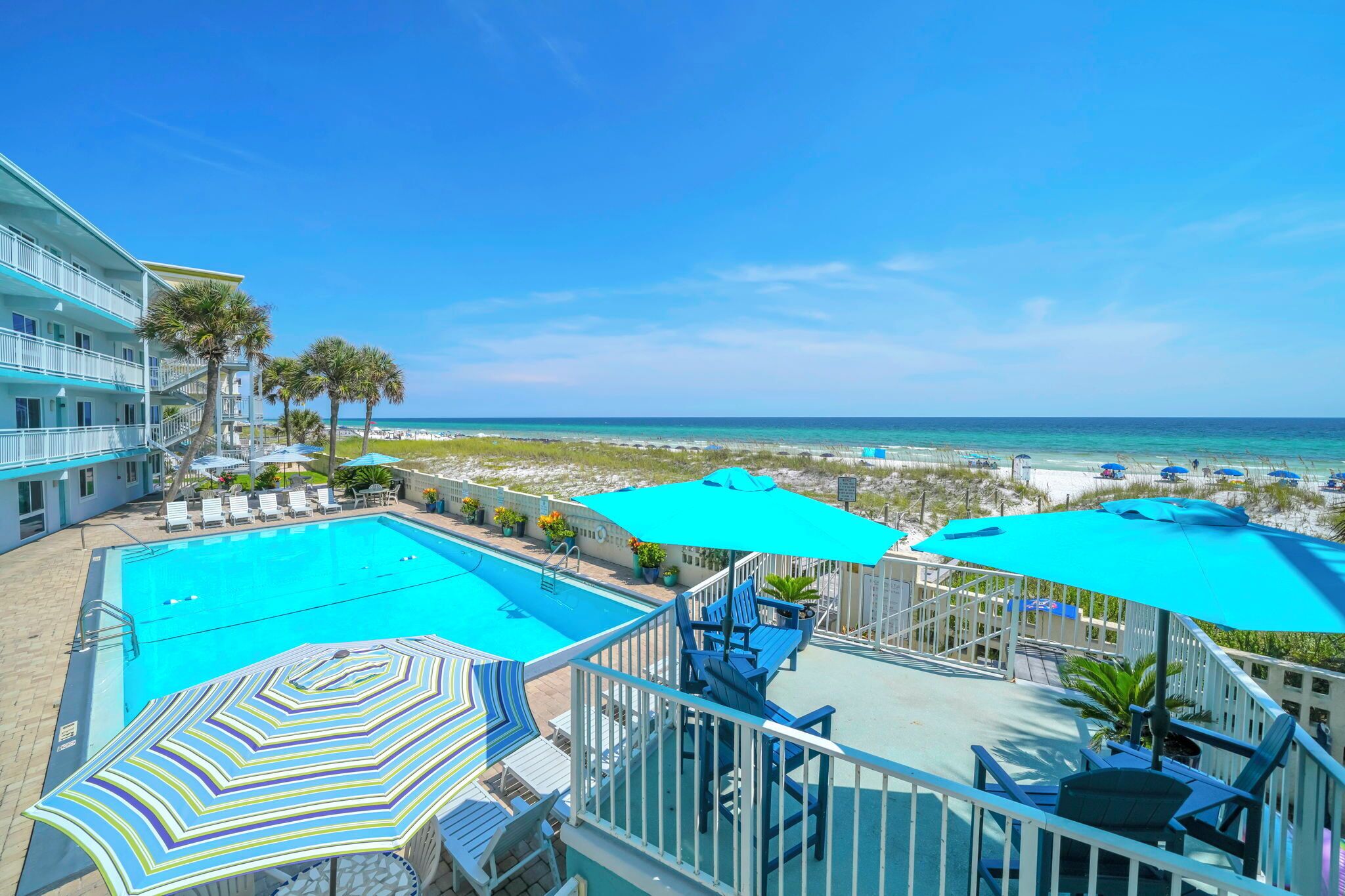 A hotel balcony with a swimming pool, beach view, and outdoor seating with umbrellas.