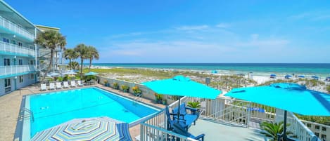 A hotel balcony with a swimming pool, beach view, and outdoor seating with umbrellas.