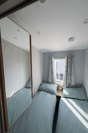 Ein kleines Zimmer mit einem Spiegelkleiderschrank, einem fenster mit Vorhängen und einem Bett mit Kopfteil.