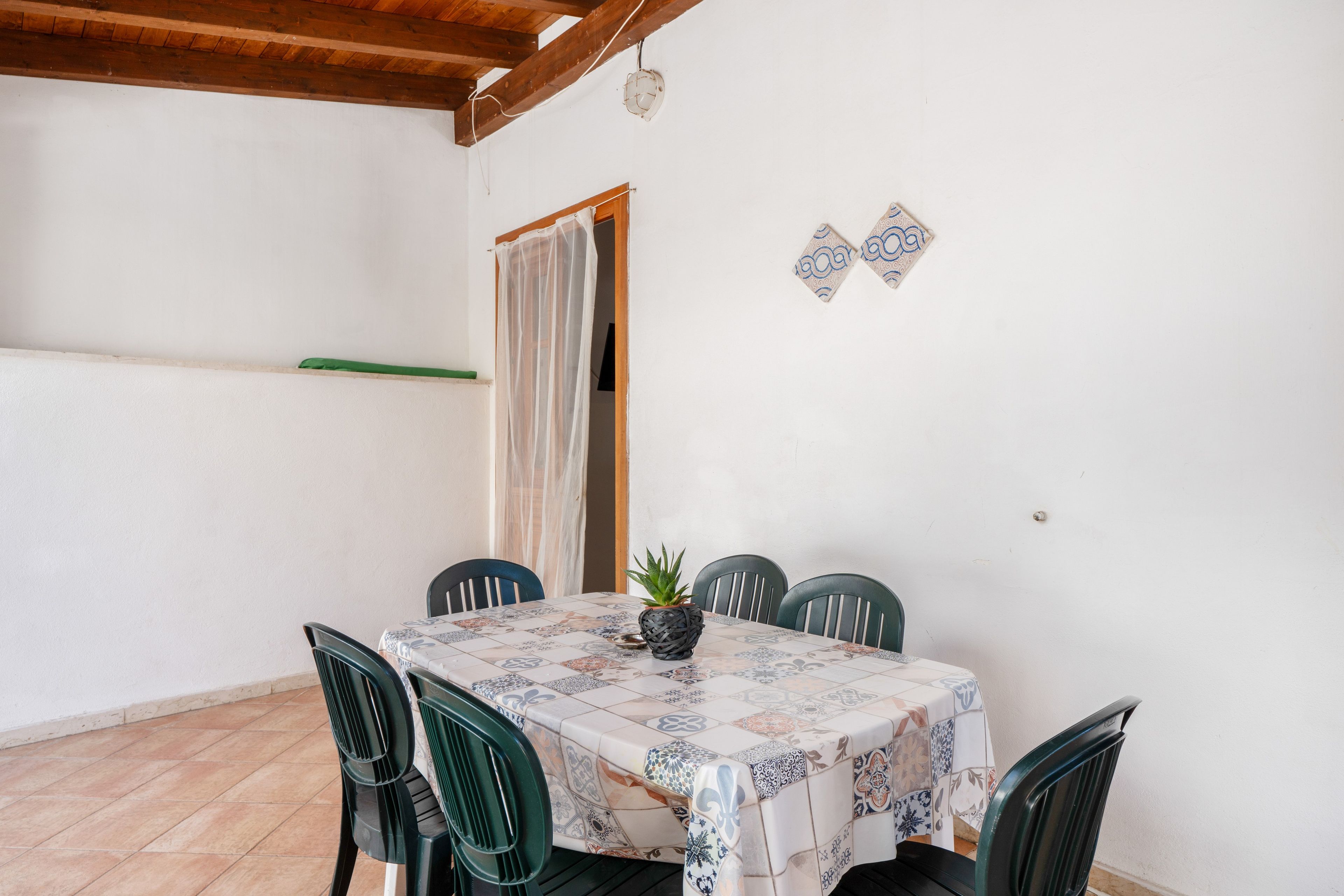 A dining area with a table covered in a patterned tablecloth, surrounded by chairs, and a potted plant on the table.