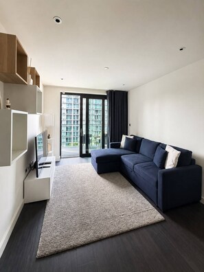 A modern living room with a dark-colored sofa, a flat-screen TV, a white entertainment unit, and a large window with a view of high-rise buildings. - Apartamento moderno a 3 min do Wembley Stadium e London Designer Outlet. (Wembley)