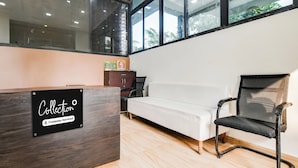 Ett väntrum med en receptionsdisk i trä, en vit soffa, en svart stol med nätmaterial och ett fönster med utsikt över grönområden.