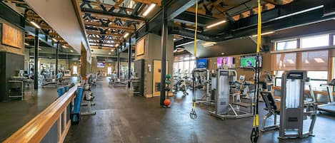 Academia com diversos equipamentos de exercícios, vigas de madeira no teto e um espelho grande na parede.