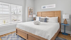 Um quarto com cama de madeira, roupa de cama branca, tapete azul e branco estampado, uma pequena mesa de cabeceira com abajur e uma janela com persianas.