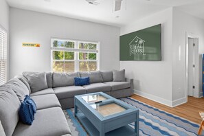 Un salon moderne avec un canapé d’angle gris, une table basse bleue et une grande fenêtre équipée de stores.