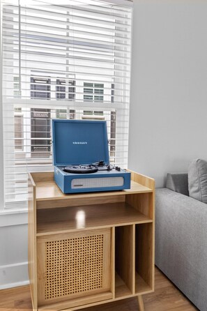 Ein Zimmer mit einem blauen Lautsprecher auf einem Holzbodenregal, einem Fenster mit Jalousien und einem grauen Sofa.