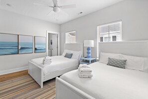 Une chambre d’hôtel avec deux lits, un ventilateur de plafond, une fenêtre avec des stores, une table de chevet avec une lampe et un mur orné de tableaux.