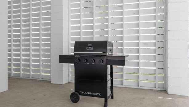Uma churrasqueira Charbroil preta com quatro queimadores e uma prateleira lateral, colocada em um espaço com paredes brancas e chão de concreto.