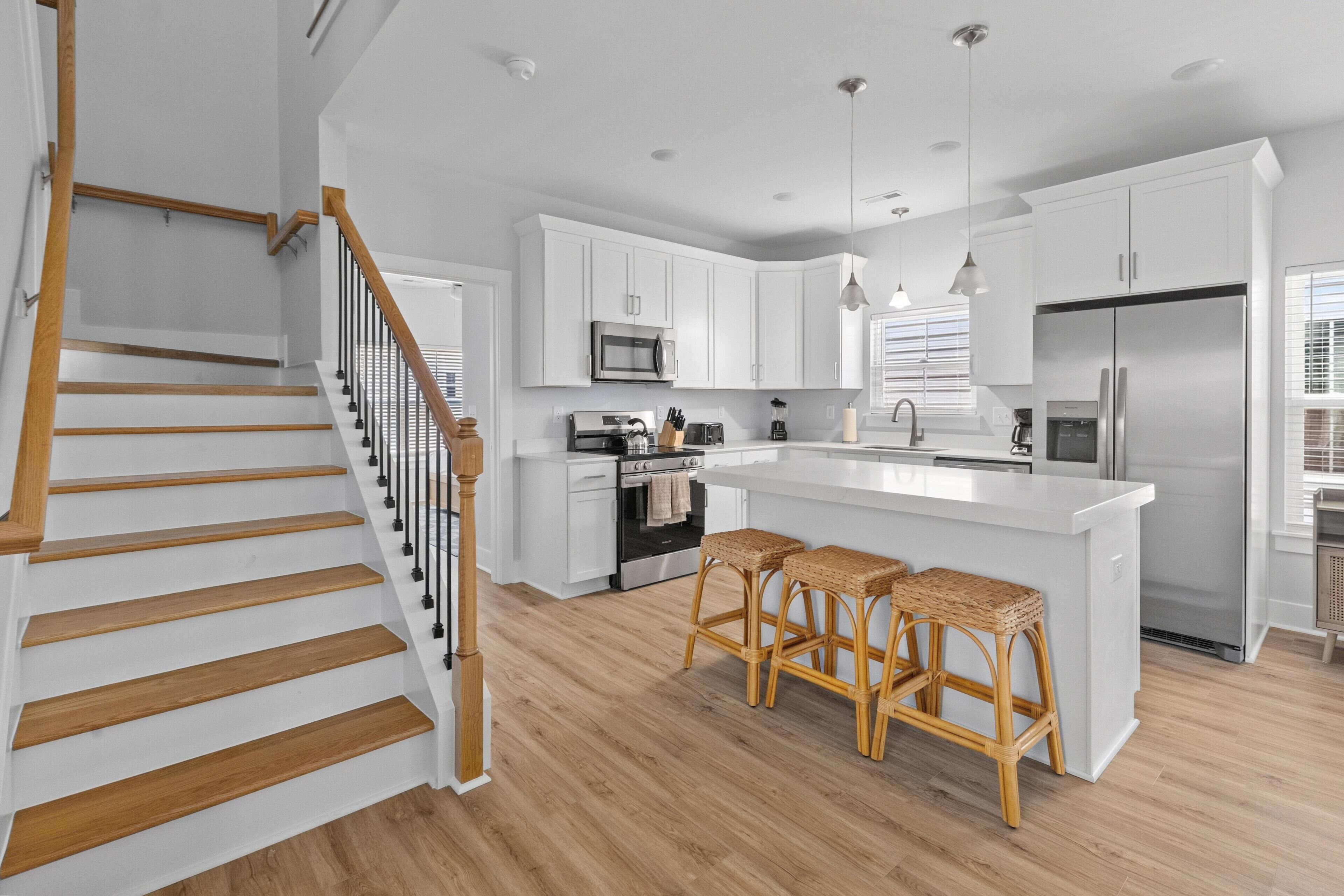 A modern kitchen with white cabinetry, a central island with bar stools, a stainless steel refrigerator, and a wooden staircase.