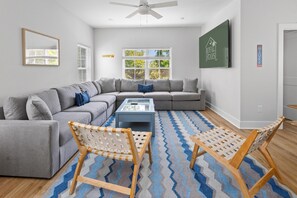 Un salon moderne avec un canapé d’angle gris, deux fauteuils en bois, une table basse en verre et un tapis bleu à motifs.