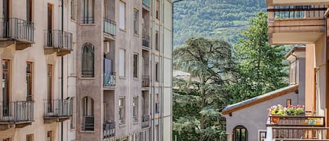 A multi-story building with balconies, a large tree in the foreground, and a mountainous backdrop.