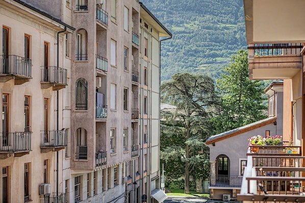A multi-story building with balconies, a large tree in the foreground, and a mountainous backdrop.