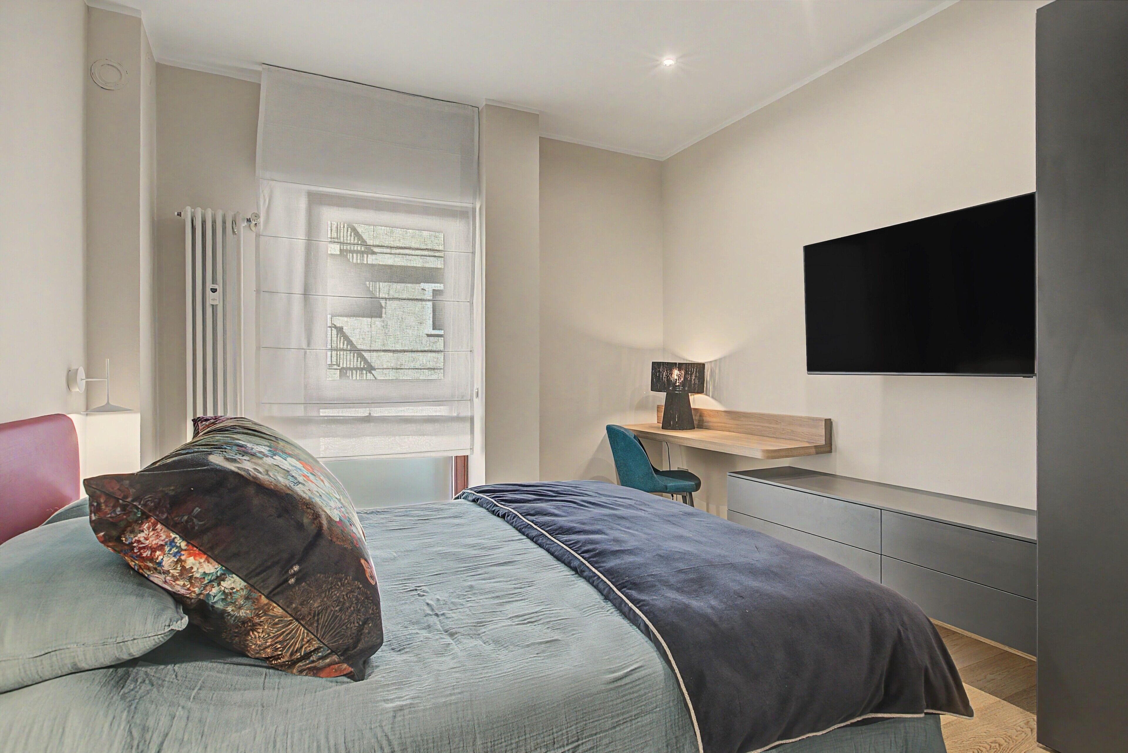 Ein Schlafzimmer mit einem Bett, einem Schreibtisch, einem Sessel, einem an der Wand befestigten Fernseher und einem Fenster mit Jalousien.
