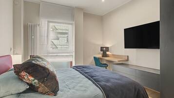Ein Schlafzimmer mit einem Bett, einem Schreibtisch, einem Sessel, einem an der Wand befestigten Fernseher und einem Fenster mit Jalousien.