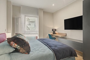 A bedroom with a bed, a desk, a chair, a TV mounted on the wall, and a window with blinds.