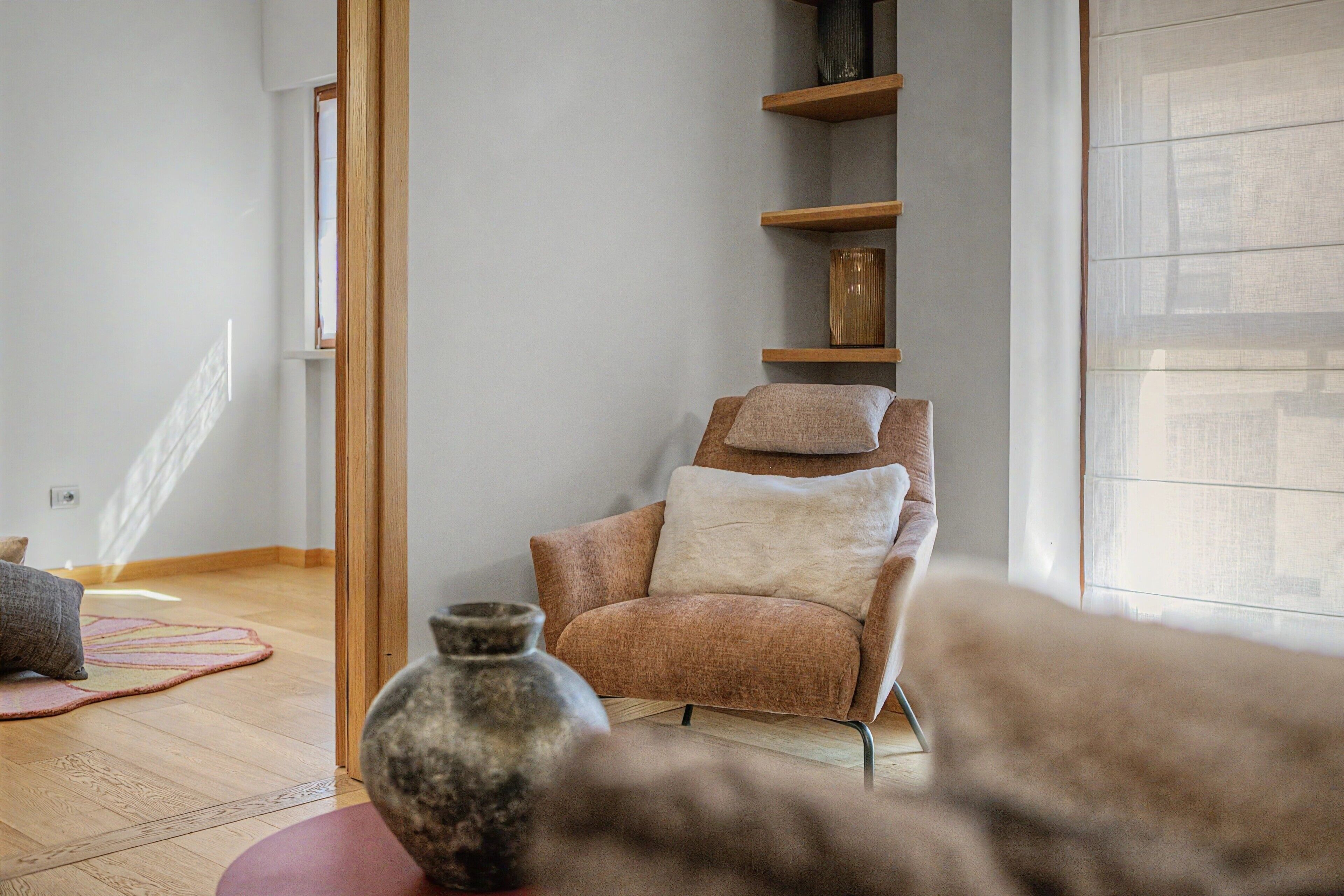 Ein Zimmer mit Holzboden, einem Sessel mit Kissen, einem Regal mit Dekoration und einem großen Fenster.