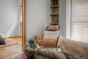 Ein Zimmer mit Holzboden, einem Sessel mit Kissen, einem Regal mit Dekoration und einem großen Fenster.