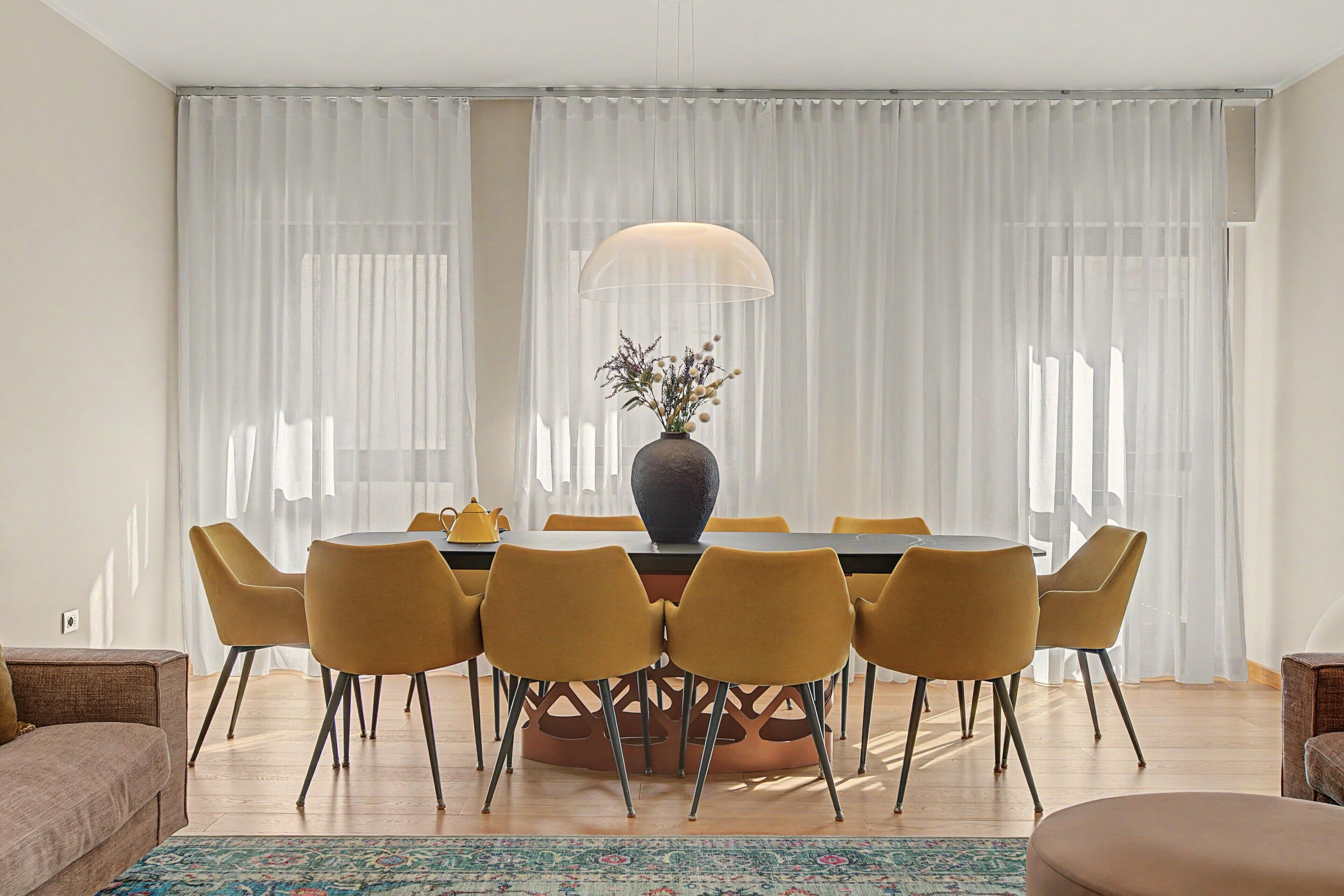 A dining area with a table, chairs, a vase with flowers, and a large window with curtains.