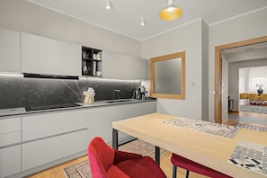 A modern kitchen with a built-in oven, a sink, and a coffee machine. There is a dining area with a table and chairs.