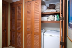 A laundry area with a washing machine, wooden louvered closets, and neatly folded towels on shelves. - Upgraded Oceanfront Condo at Mauna Lani Terrace (Kamuela)