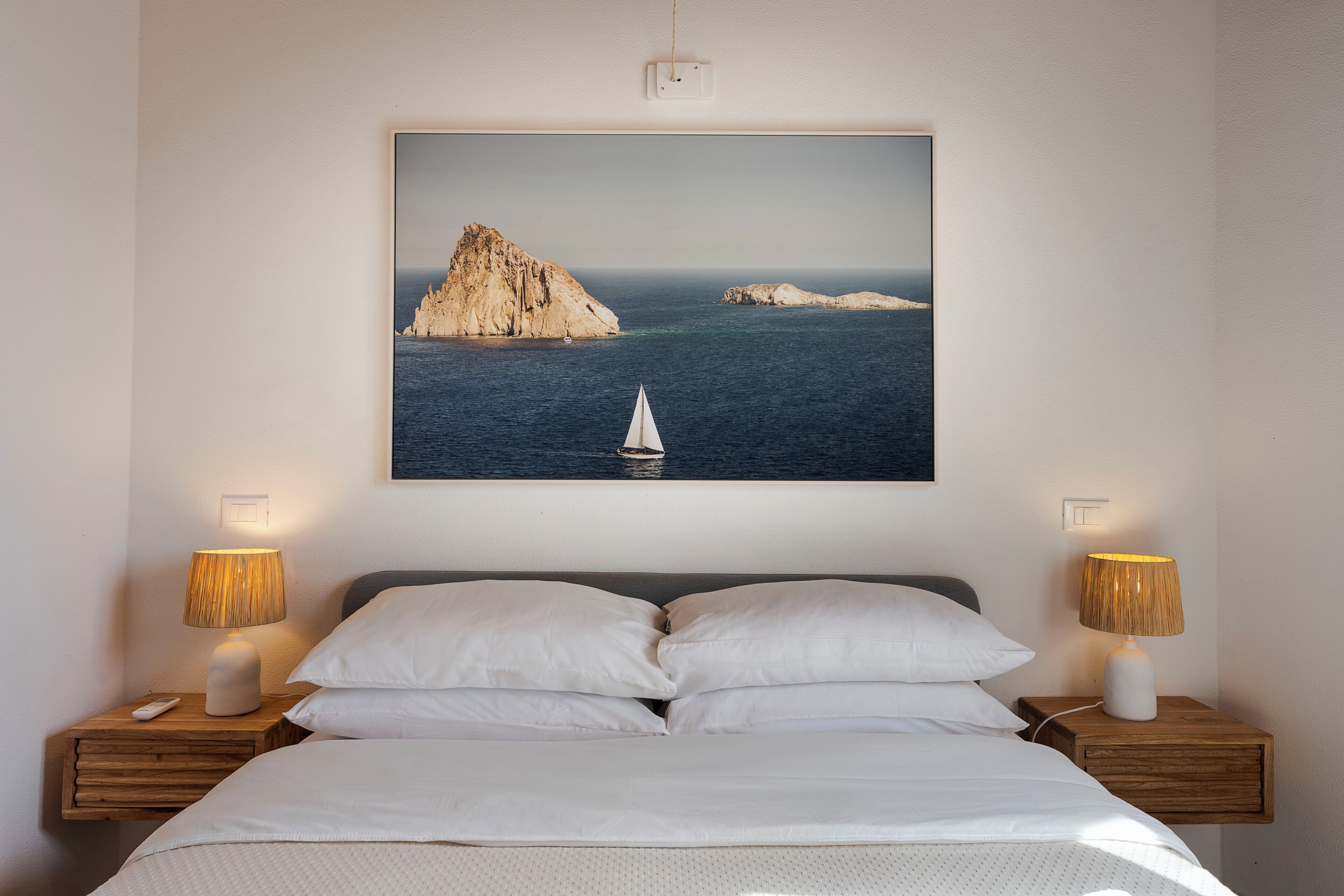 Ein Bett mit weißen Leinen, zwei Nachttischlampen und ein gerahmtes Bild einer Küstenlandschaft mit einem Segelboot.