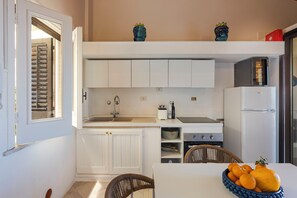 Una cocina funcional con armarios blancos, un lavabo, una estufa y un refrigerador. Hay jarrones decorativos en la encimera y una silla de mimbre con un cuenco de fruta sobre la mesa.