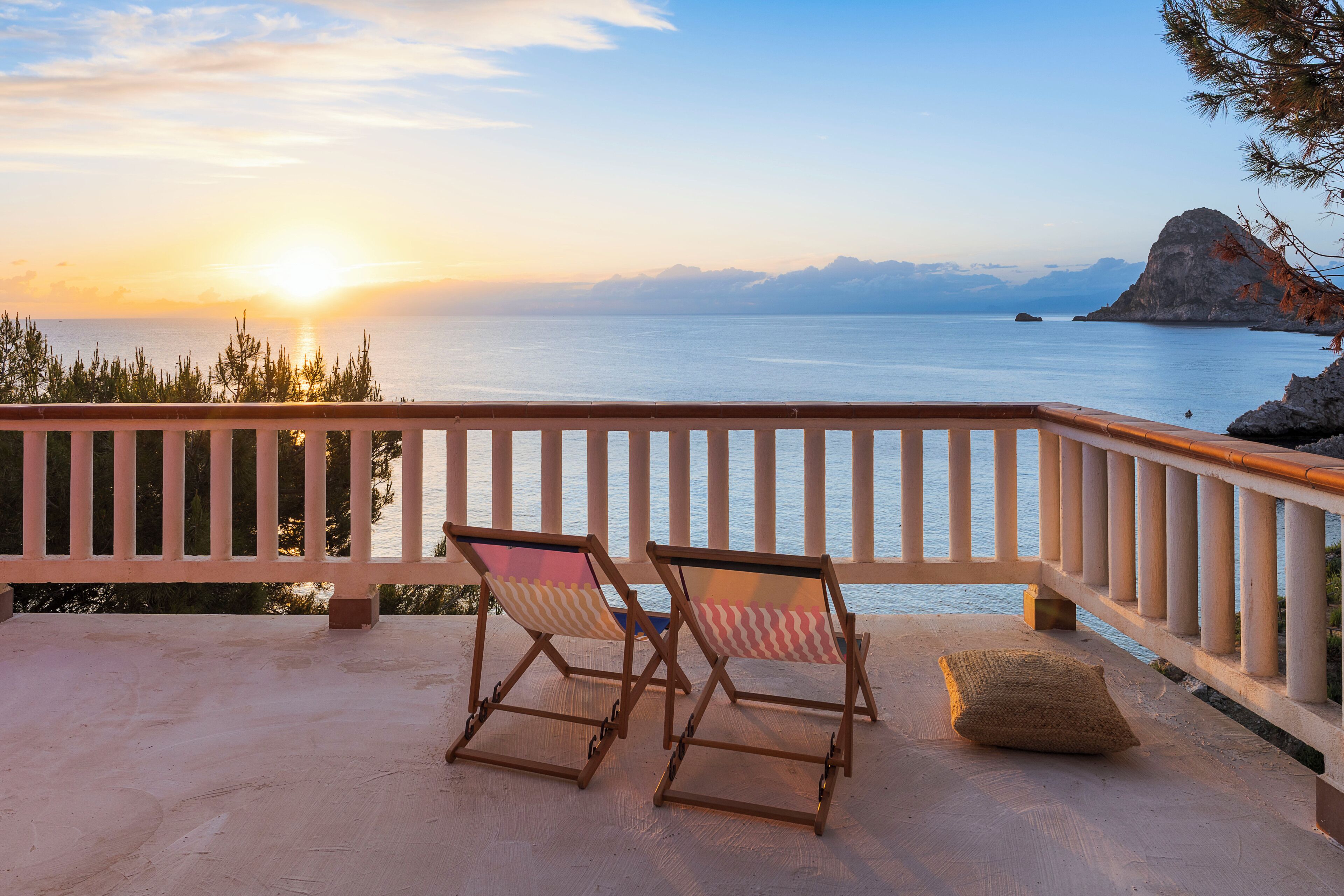 A balcony with two wooden chairs and a cushion, overlooking a calm sea and distant mountains during sunset.
