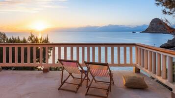 A balcony with two wooden chairs and a cushion, overlooking a calm sea and distant mountains during sunset.
