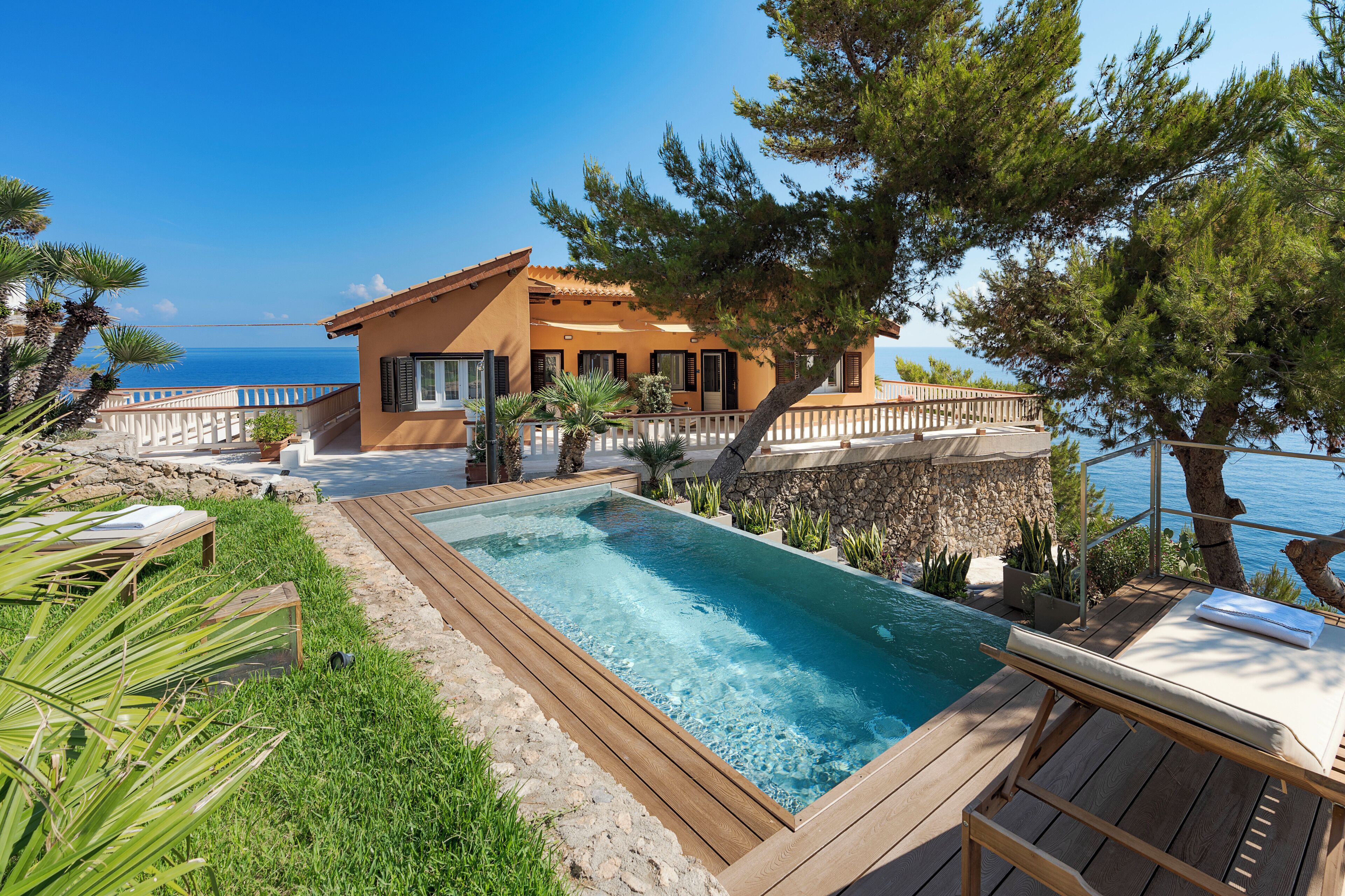 A pool with a wooden deck, a house with a tiled roof, and a view of the sea.
