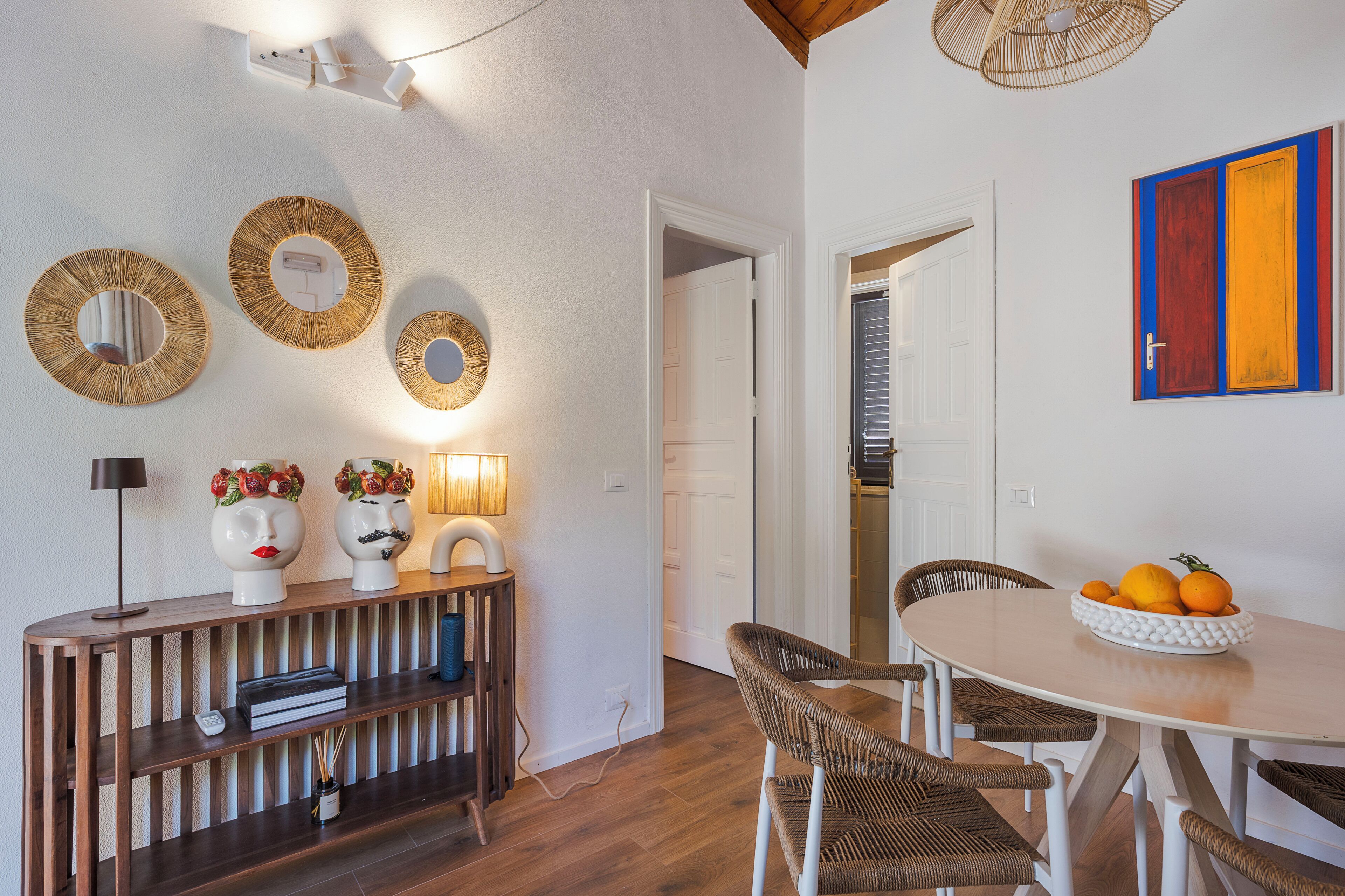 A dining area with a round table, wicker chairs, a wooden console table with decorative items, and wall-mounted mirrors.
