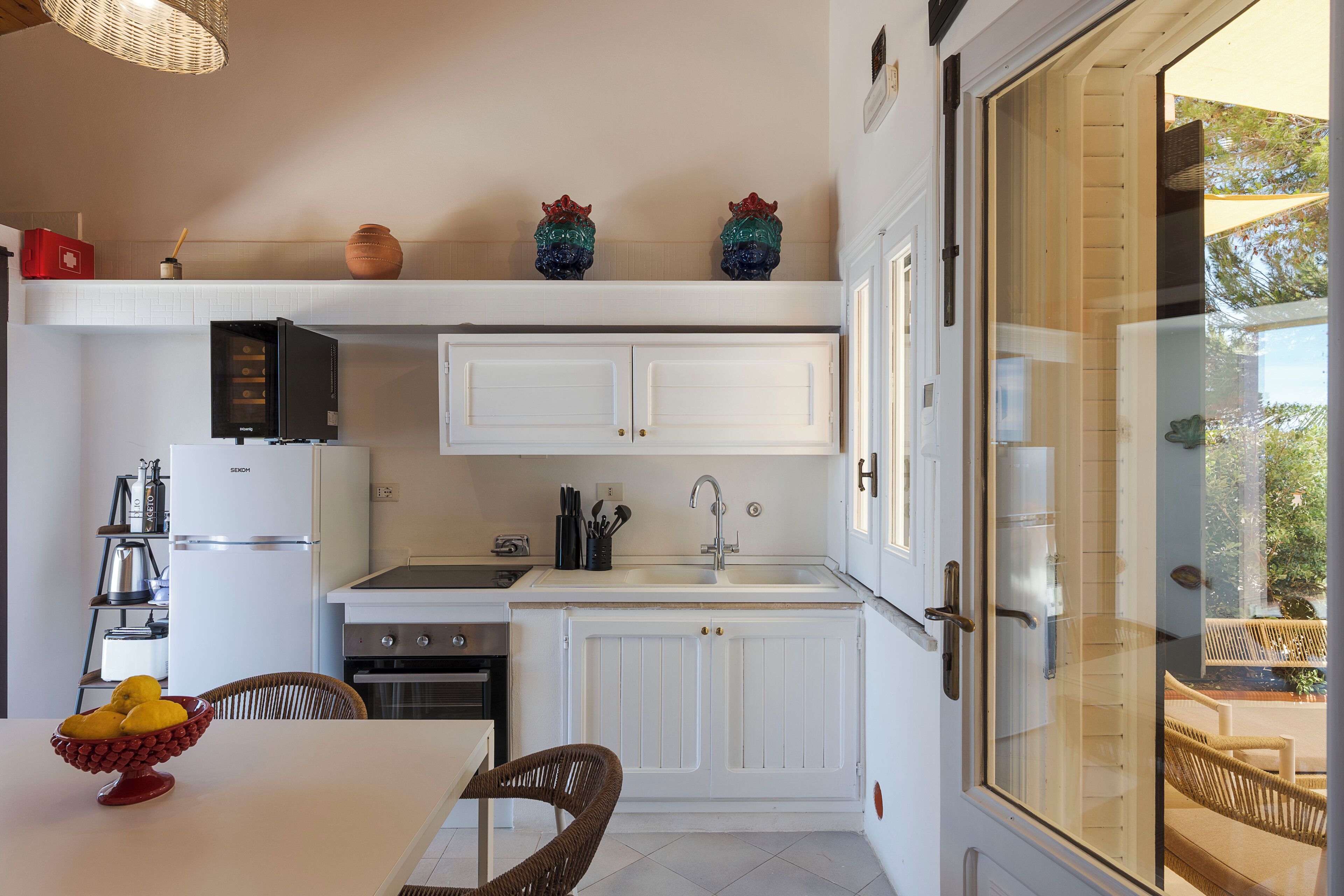 A compact kitchen with white cabinetry, a built-in stove, and a dining area with a wicker chair.