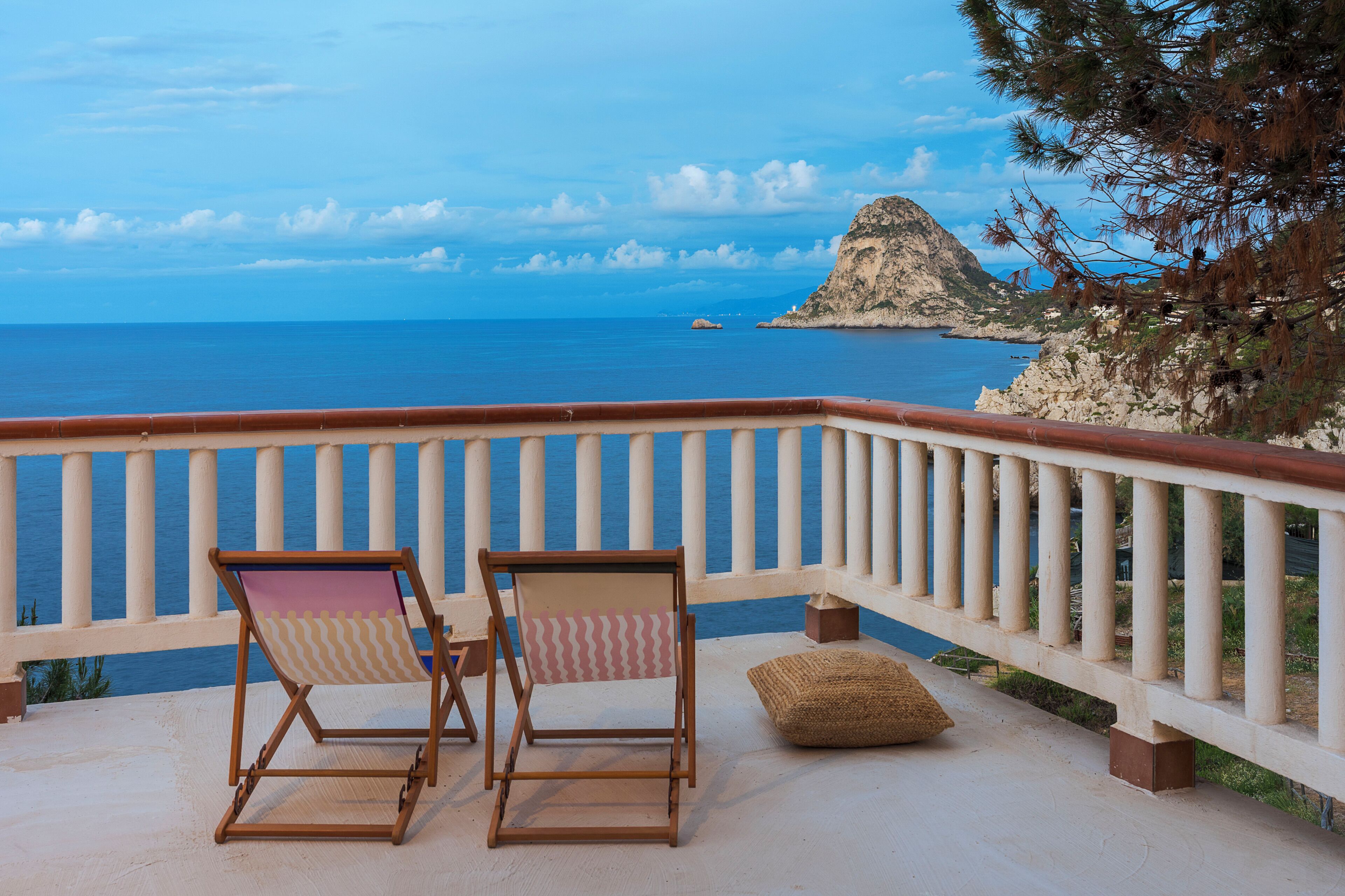 A balcony with two wooden chairs and a cushion, overlooking a coastal view with a rocky outcrop and the sea.