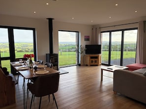 A spacious living room with a fireplace, a dining area set for a meal, and large windows offering a view of the countryside. - High View Barn (Launceston)