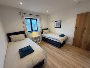 A bedroom with two beds, a window with curtains, a bedside table with a lamp, and a wooden wardrobe. - High View Barn (Launceston)