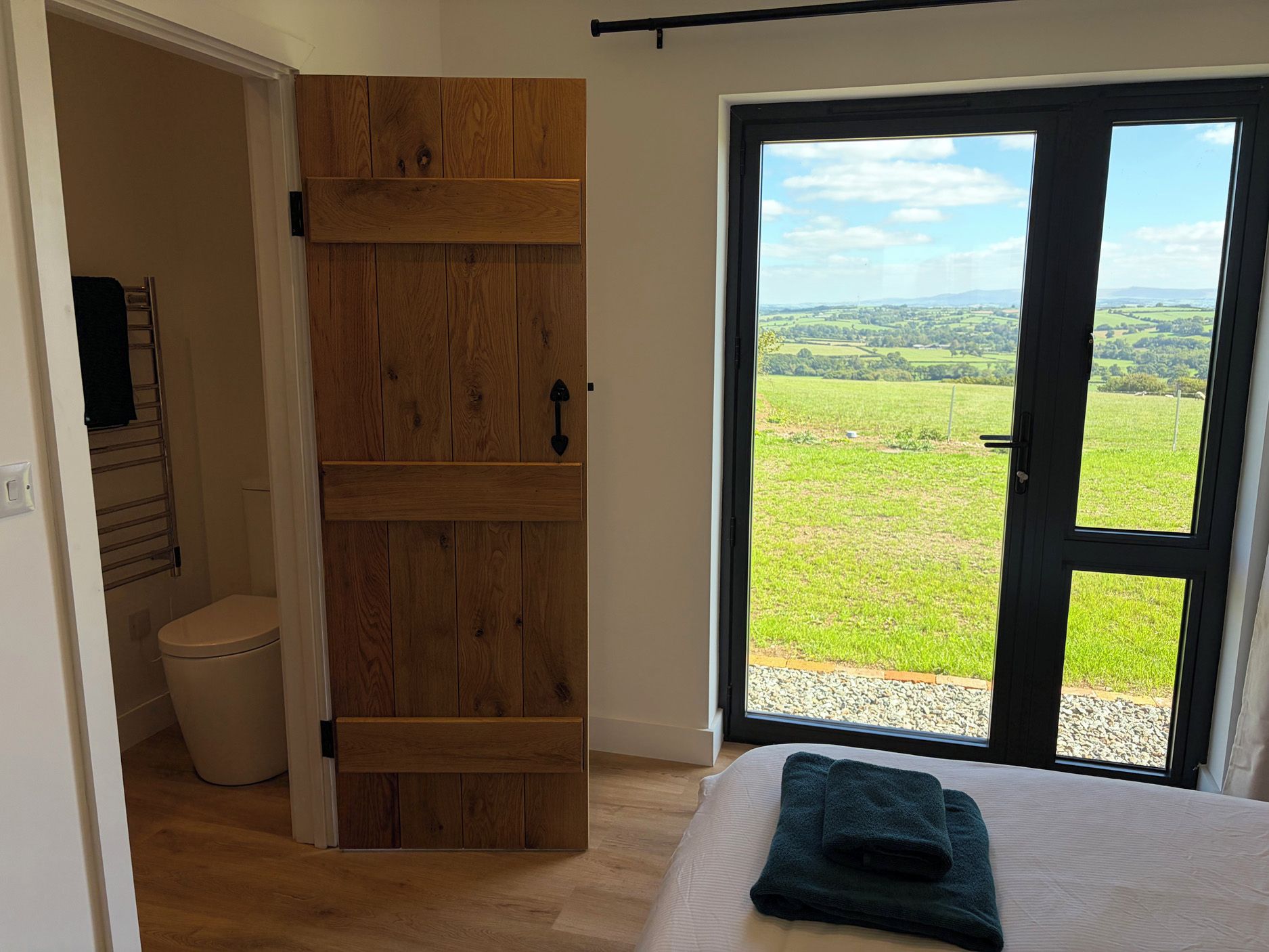 Een slaapkamer met een houten deur naar een badkamer, een bed met een opgevouwen handdoek en uitzicht op een groen landschap door een glazen deur.