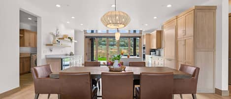 A modern kitchen with wooden cabinets, a dining table with chairs, and a large window offering a view of greenery.