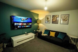 A living room with a flat-screen TV mounted on the wall, a black sofa with colorful cushions, a white sideboard, and a small round table. - 3-Bed Serviced Home | Ensuite | Work Desk| Bilston (West Midlands)
